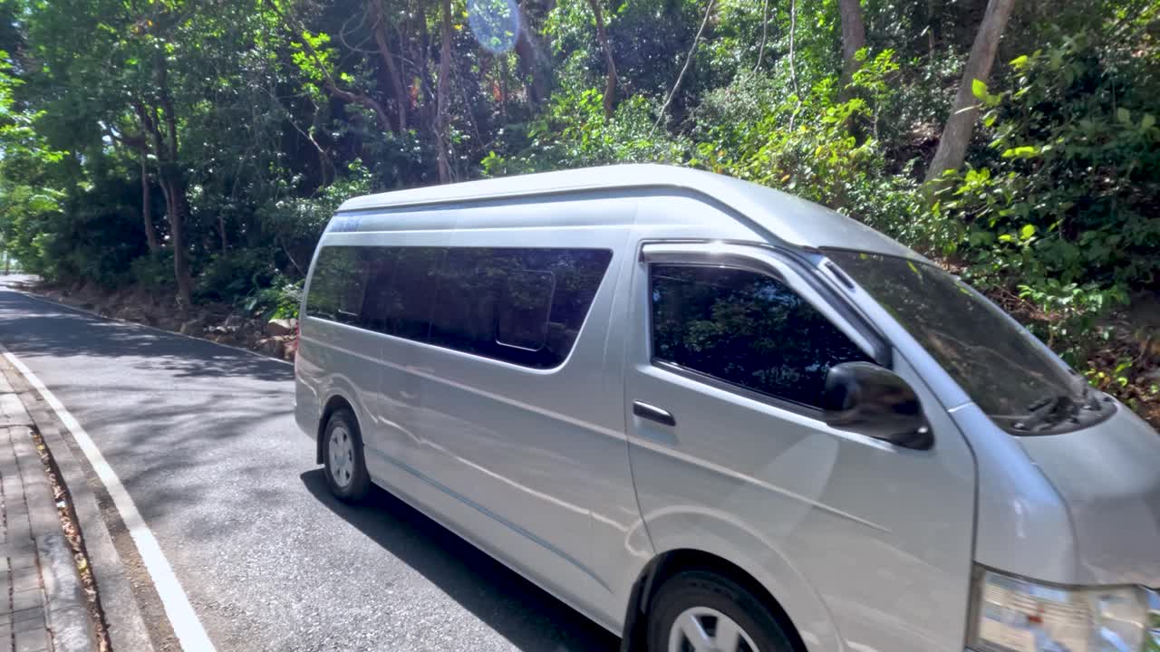 White passenger van drives along winding forest road in bright daylight, lush tropical surroundings visible