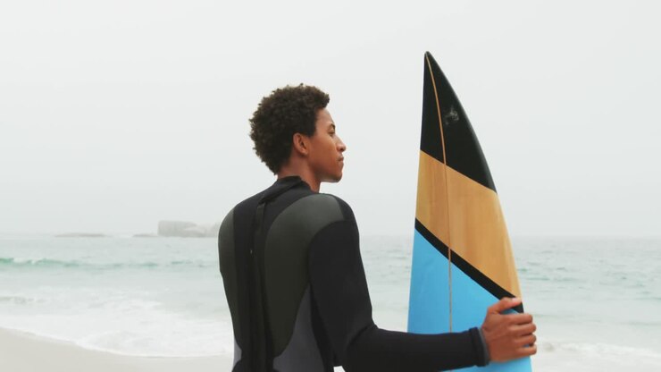 Vista posteriore di surfista afroamericano in piedi con la tavola da surf sulla spiaggia 4k