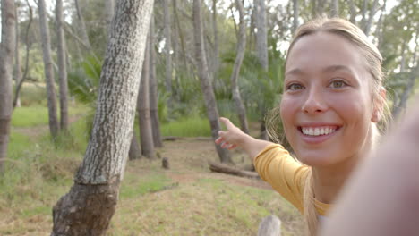 Young-woman-with-blonde-hair-smiles-at-the-camera,-extending-her-arm-towards-the-trees-with-copy-spa