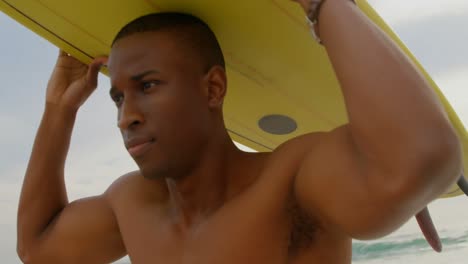 vista frontal de un surfista afroamericano que lleva una tabla de surf en la cabeza en la playa 4k