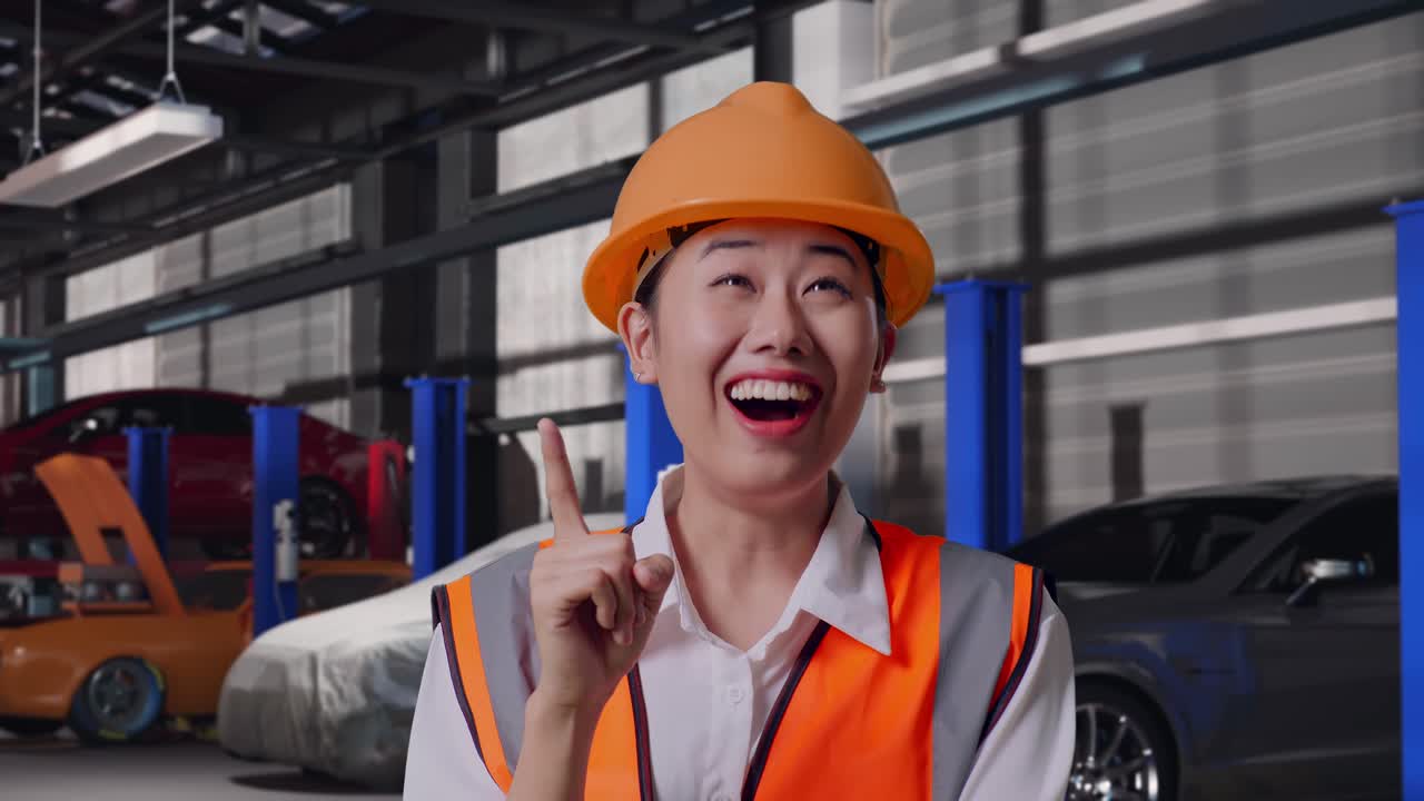 Nærbilde av asiatisk kvinnelig ingeniør med vernehjelm som tenker og ser seg rundt, og deretter løfter pekefingeren mens hun står i et bilverksted