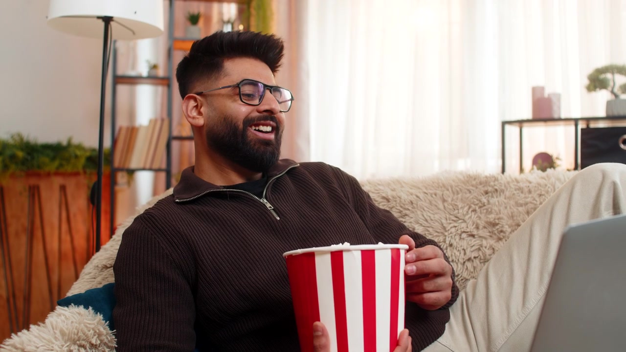 Uomo indiano che guarda un film sul portatile e mangia popcorn a casa sorridendo e godendosi il tempo libero