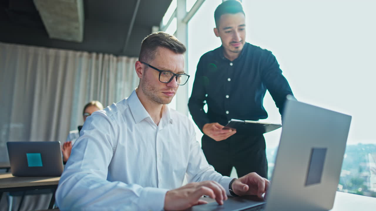 Serious mid-aged man wearing white shirt and glasses works on computer. His colleague standing beside with documents in his hands. Female at desk at backdrop.