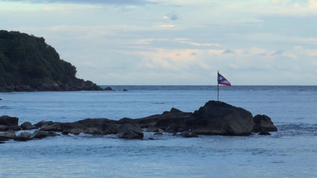 Vista panoramica della bandiera portoricana che sventola su una costa rocciosa vicino a onde oceaniche blu che si infrangono, sotto un cielo luminoso a Porto Rico.