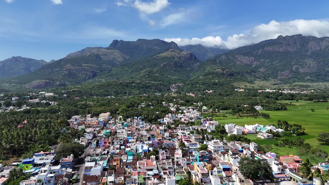 Aerial view of a vibrant South Indian town nestled directly at the foothills of a majestic mountain range. The foreground features a dense cluster of colorful houses and a vivid green paddy field