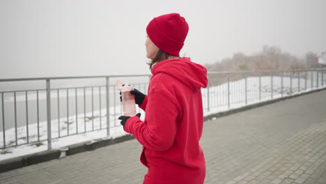 dama con capucha roja y gorra beba agua de botella rosa al aire libre durante la caminata de invierno, dejando caer la botella en un entorno sereno con camino cubierto de nieve, barandilla de hierro y fondo atmosférico brumoso