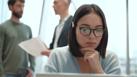 an young woman working with a laptop while two men talk in the background behind her 1