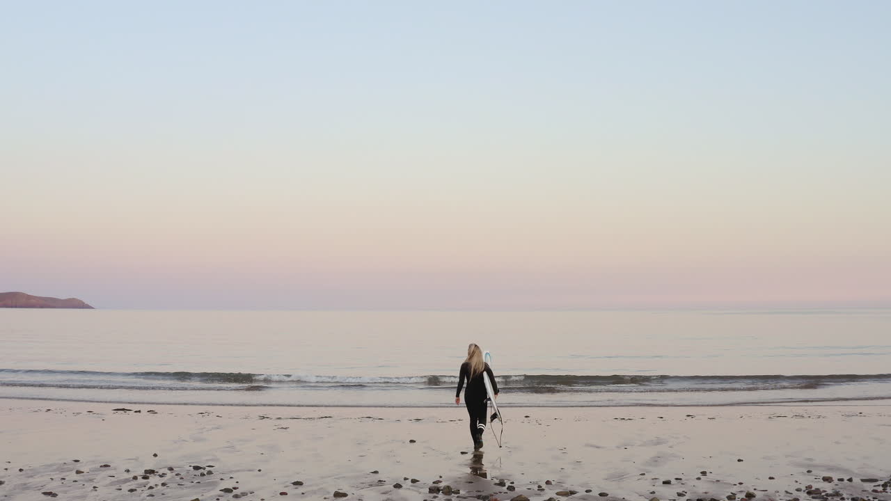 바다로 걸어가는 서프보드를 들고 있는 웨이트슈트를 입은 여성의 뒷면