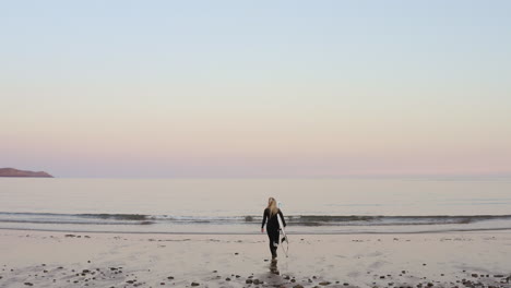 Rear-View-Of-Woman-Wearing-Wetsuit-Carrying-Surfboard-Walking-Into--Sea