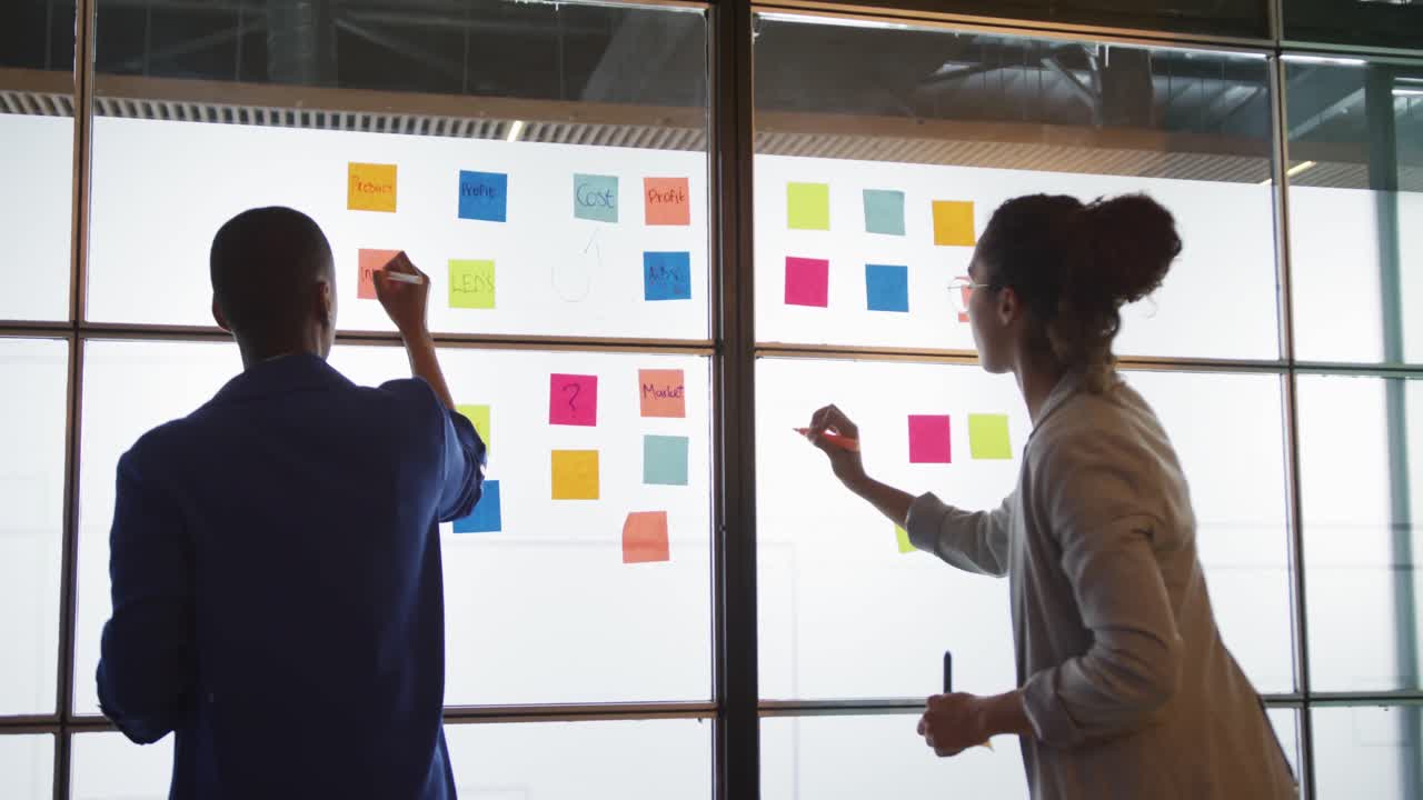 Diverse female creative businesswomen writing on memo notes, brainstorming on glass wall in office