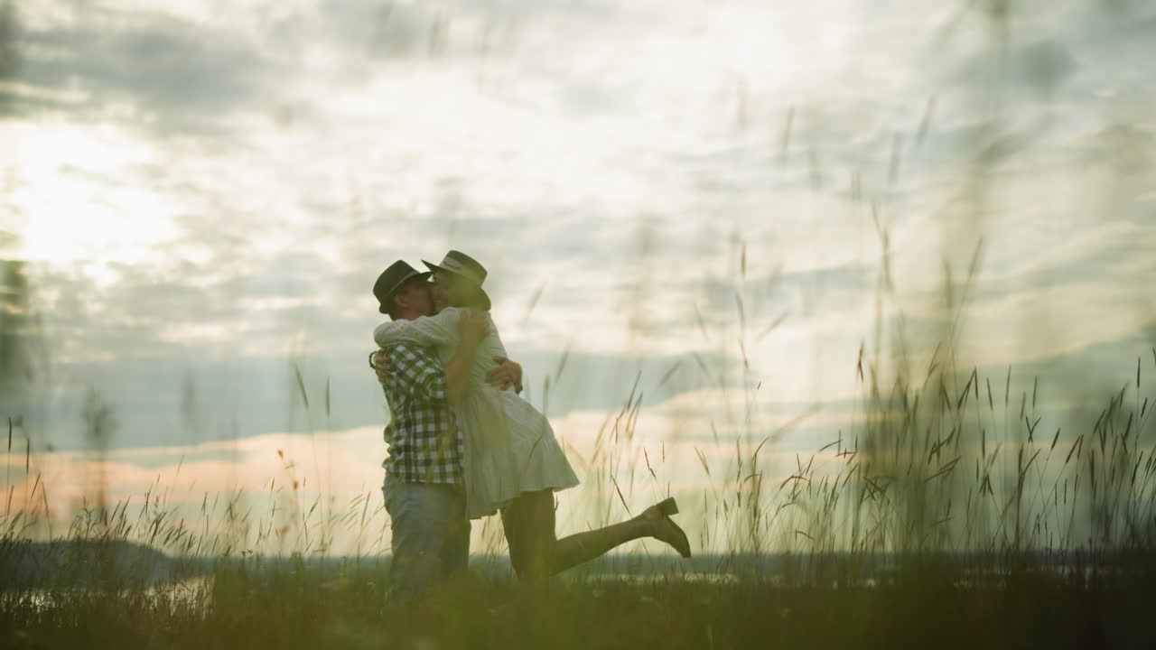 A loving couple shares a tender moment as the man gently kisses the woman's cheek in a peaceful grassy field under a cloudy sky at sunset. She wears a white dress, and he wears a checkered shirt