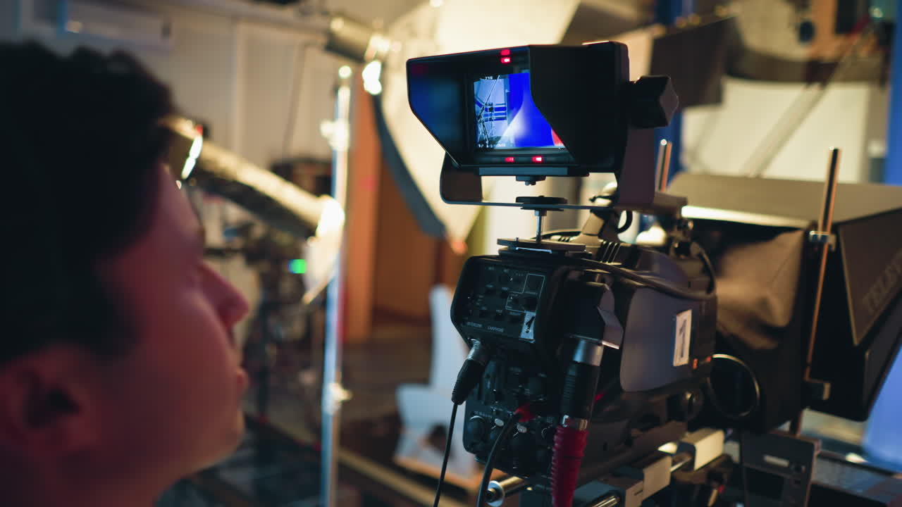 Cameraman views professional studio camera screen showing female presenter in red outfit on blue screen stage during live recording session, surrounded by production lighting, teleprompter, and technical gear