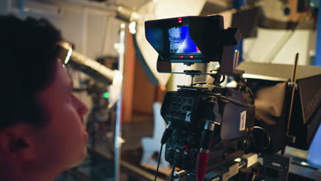 cameraman views professional studio camera screen showing female presenter in red outfit on blue screen stage during live recording session, surrounded by production lighting, teleprompter, and technical gear
