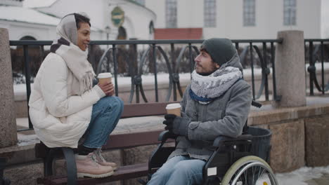 muslim woman and her disabled friend in wheelchair drinking takeaway coffe on a bench in city in winter