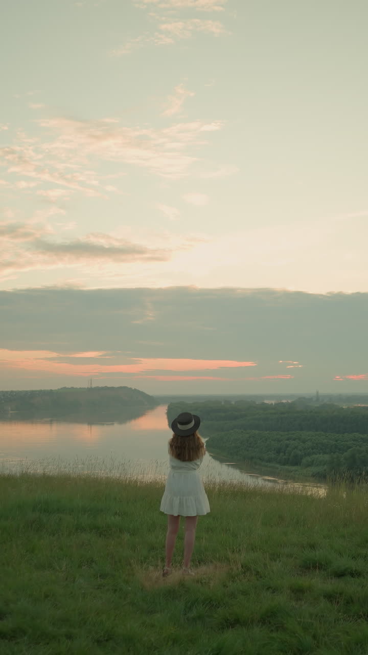 A woman in a white dress and hat stands thoughtfully in a grassy field overlooking a tranquil lake at sunset. She gazes into the distance, capturing a peaceful, reflective moment as the sun sets