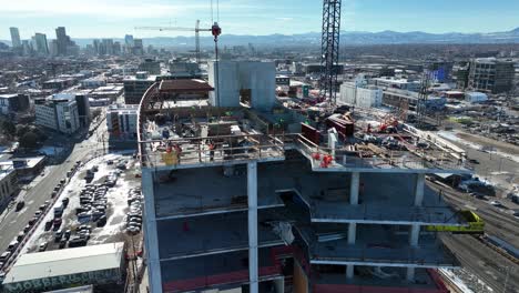 drone volando alrededor de edificios de varios pisos en construcción y trabajadores que trabajan en el techo con vistas a la ciudad en el fondo, denver