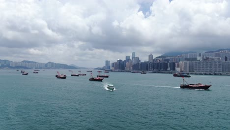 Convoy-De-Barcos-De-Pesca-Locales-Que-Causan-En-La-Bahía-Victoria-De-Hong-Kong,-Con-El-Horizonte-De-La-Ciudad-En-El-Horizonte,-Vista-Aérea