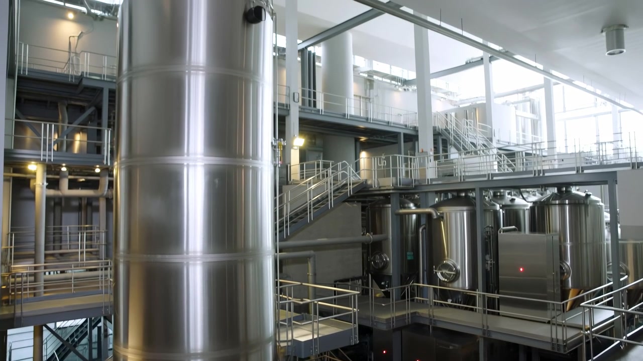 Large reflective metal tank in the foreground of a clean industrial facility, with rows of stainless steel vessels, pipes, stairs, railings, and elevated