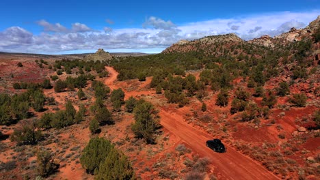 Hermosa-Antena-De-Un-Auto-Negro-Conduciendo-A-Través-Del-Desierto-Con-Pocos-árboles-Alrededor-En-Un-Terreno-Montañoso