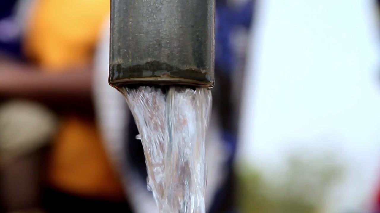 Una vista ravvicinata di acqua limpida che scorre costantemente da un tubo di metallo scuro, con uno sfondo sfocato che suggerisce un ambiente esterno
