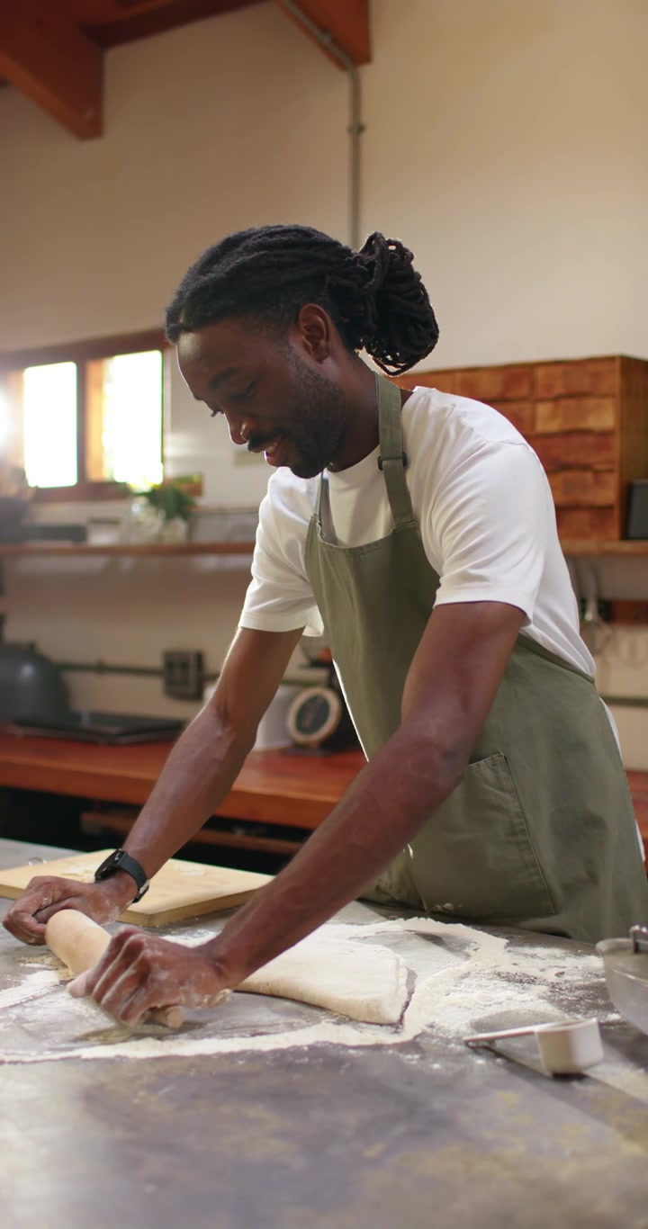 Vertical video: African American man in apron laying rolling pin, rolling dough for baking at bench