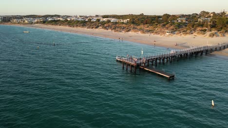 Aerial-orbiting-shot-of-people-on-jetty-and-resting-tourist-at-sandy-beach-in-Perth-City-during-golden-sunset