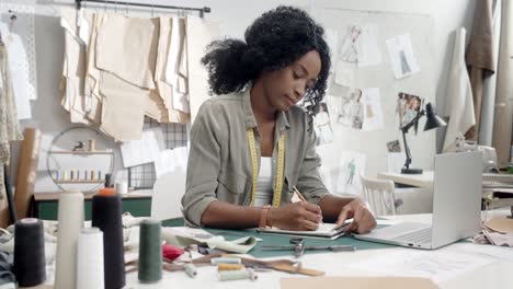 woman clothes designer sitting and working at the laptop computer, drawing outlines in her notebook and smiling to the camera in her studio