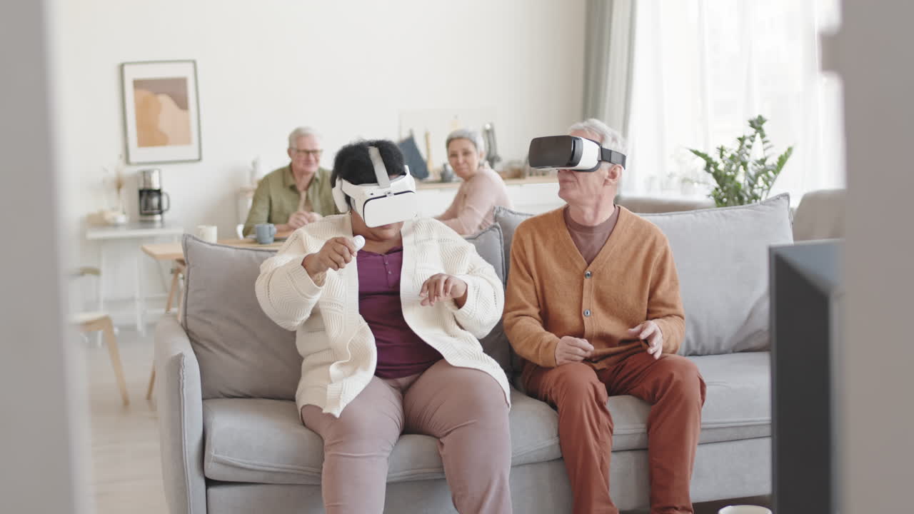Piano americano di uomo e donna divertiti che indossano visori per la realtà virtuale, seduti sul divano in salotto, muovendo la testa, sorridendo