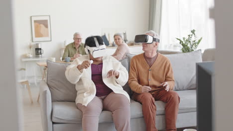 medium long of amused man and woman wearing virtual reality headsets, sitting on couch in living room, moving heads around, smiling