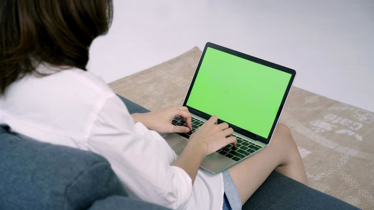 joven asiática usando un dispositivo portátil con pantalla verde. mujer asiática trabajando portátil mientras está sentada en el sofá en la sala de estar en casa. llave de croma.