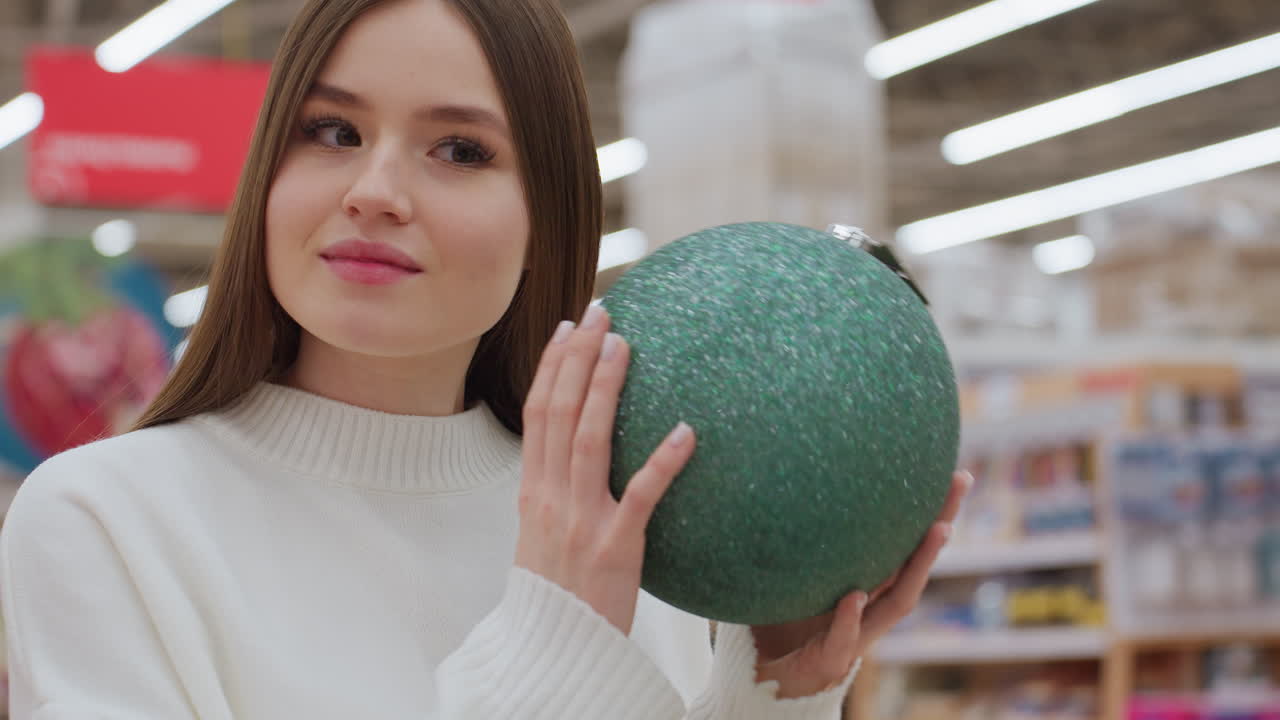 Elegant lady holding green shimmering ball decor, shaking it with a warm smile in a well-lit decor store, surrounded by beautiful Christmas decorations and shoppers in the background