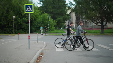 friends walking side by side with their bicycles across a pedestrian crossing, with a blurred view of two people walking in the background, a parked car is visible, and there are trees visible around