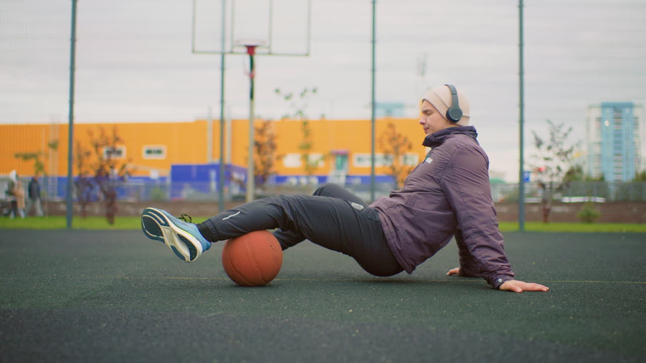 Defaticamento su un campo da basket all'aperto con persona di razza sconosciuta seduta sull'asfalto con un pallone da basket sotto le gambe, che indossa una giacca e cuffie, appoggiata all'anello urbano con le mani, recinzione e skyline