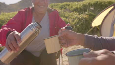 happy senior biracial couple sitting at tent in mountains and drinking coffee, in slow motion