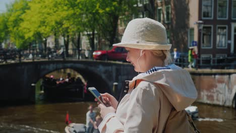 a tourist woman uses a smartphone in the center of amsterdam. in the background, the intensive movement of boats, pedestrians and cyclists