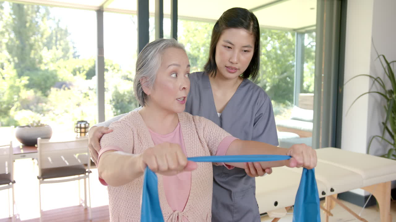 Senior asian woman exercising with resistance band, assisted by physiotherapist at home