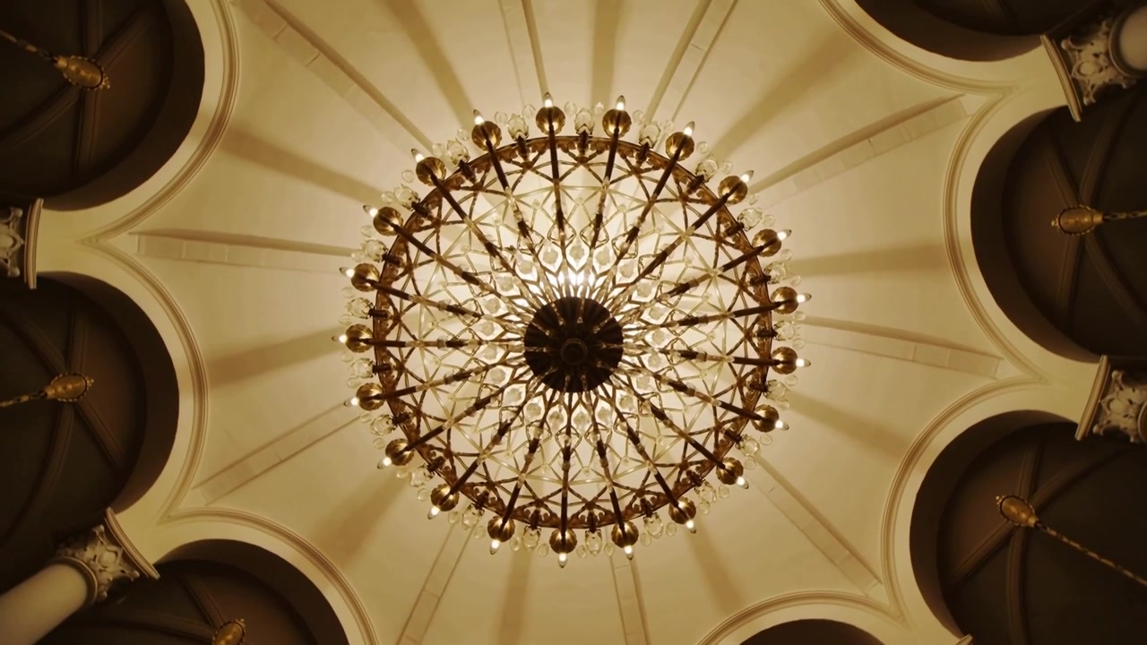 Telecamera che si inclina, catturando un grande lampadario circolare in una sala grande, rivelando costoloni e archi ornati.