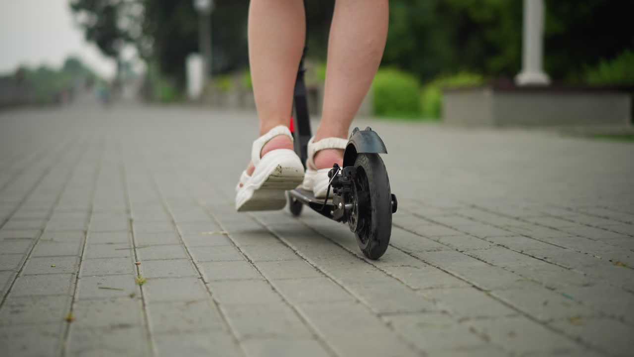 Close-up of a woman riding a black scooter on a paved walkway, her legs are visible as she moves forward, with a child riding a green scooter in the background fast pass her