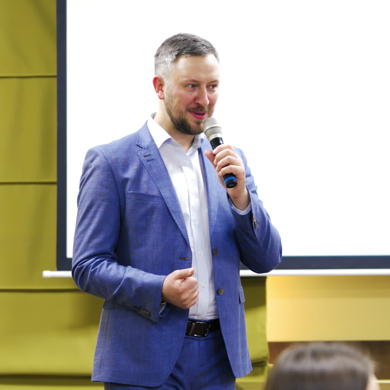 Business leader male conducts a lecture with a smile on his face during the conference. Man in stylish blue suit with microphone speaking on the background of white screen.