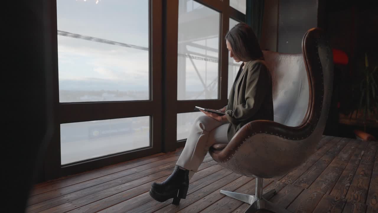 revelación de la hembra sentarse en la silla de huevo de cuero retro y sostener la tableta cerca de una amplia ventana