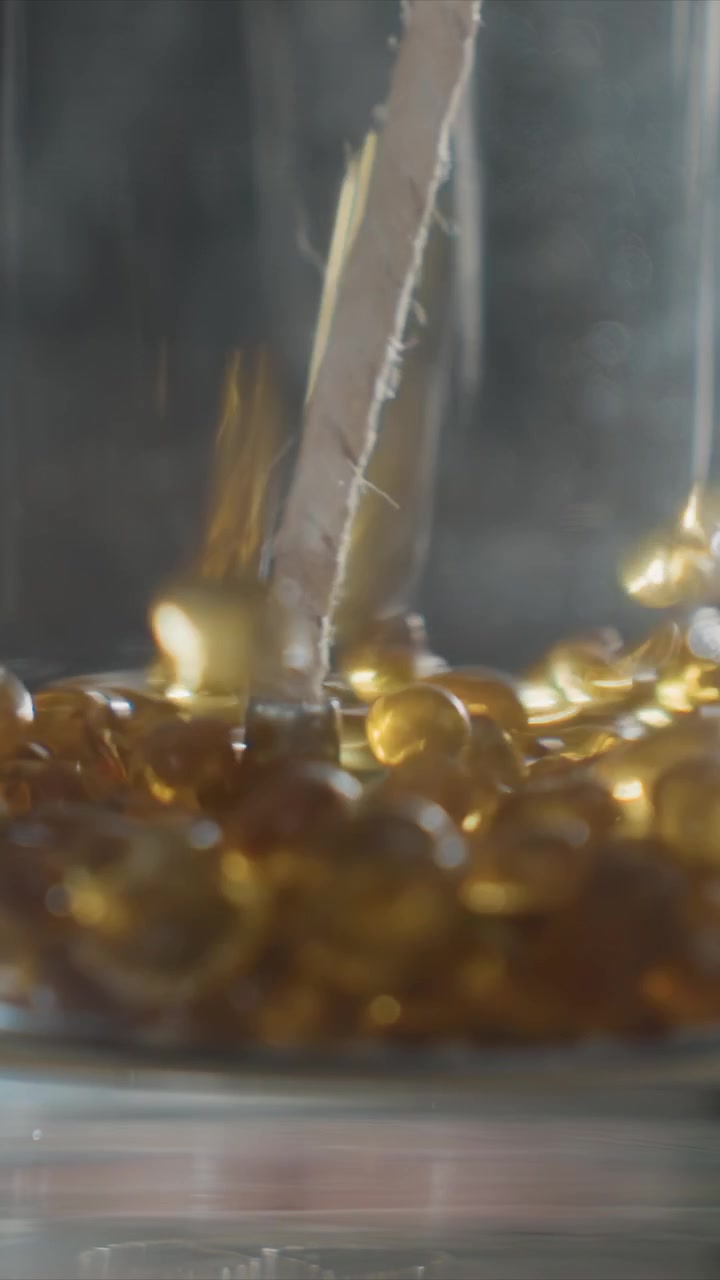 Primo piano di goccioline di liquido dorato che si formano attorno allo stoppino di una candela in un contenitore di vetro. Perle di cera si accumulano alla base mentre la luce si riflette sulle loro superfici lisce creando morbidi riflessi luminosi.