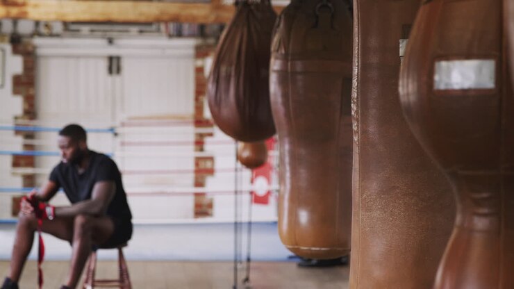 Pugile che si allena in palestra, si fascia le mani seduto accanto al ring e ai sacchi da boxe.