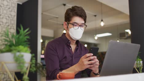 Hombre-Asiático-Con-Mascarilla-Usando-Un-Teléfono-Inteligente-Mientras-Está-Sentado-En-Un-Café