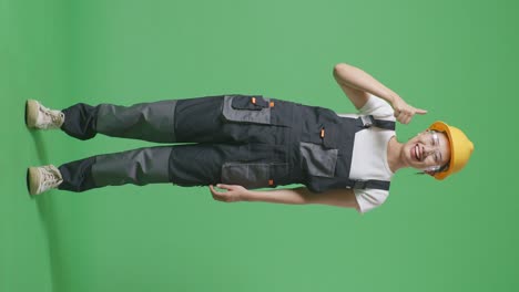 cuerpo lleno de mujer asiática trabajadora con gafas de protección y casco de seguridad sonriendo y mostrando pulgares hacia arriba gesto mientras está de pie en el estudio de fondo de la pantalla verde