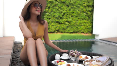 woman wearing hat, sunglasses and swimsuit sitting by plunge pool with breakfast on floating tray