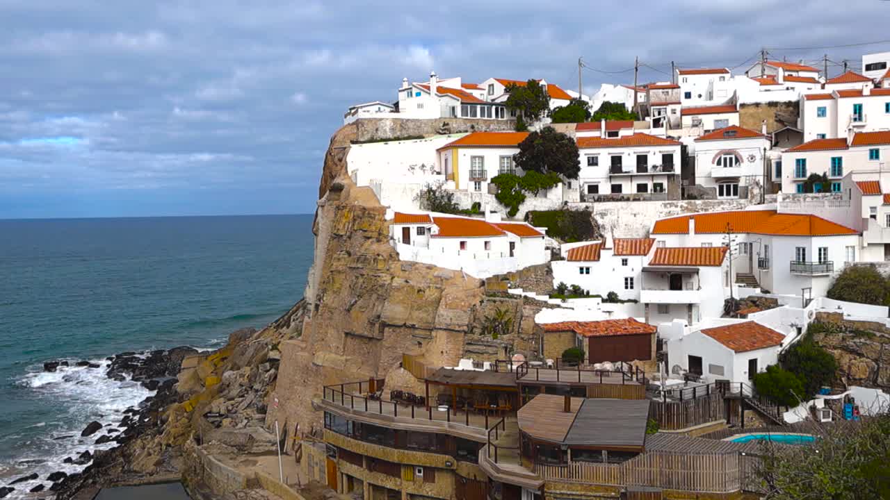 Una vista panorámica lenta desde un punto elevado revela el pueblo costero de Azenhas do Mar en Sintra. Casas blancas con tejados rojos encaramadas sobre el océano. Olas del Atlántico rodando hacia acantilados escarpados