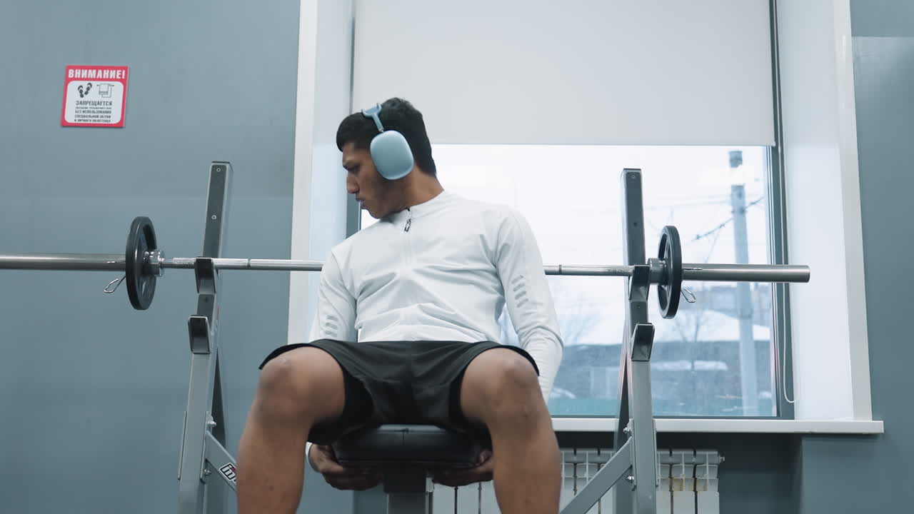 Young student carrying metal weight plate across gym floor to place onto barbell rack beside window showing residential building outside