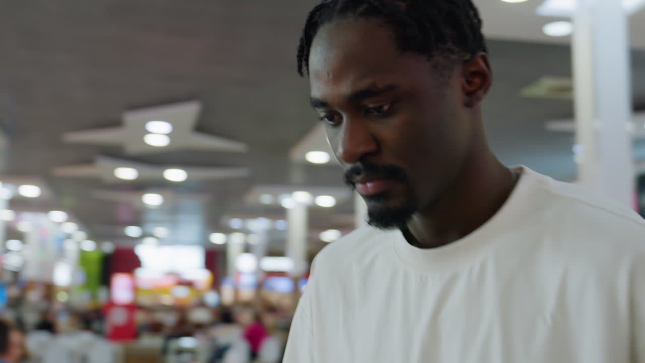 Close up of young african man wearing white shirt sitting in cafe dropping tray with snack and drink as he bends forward with focused expression under indoor lighting and blurred background