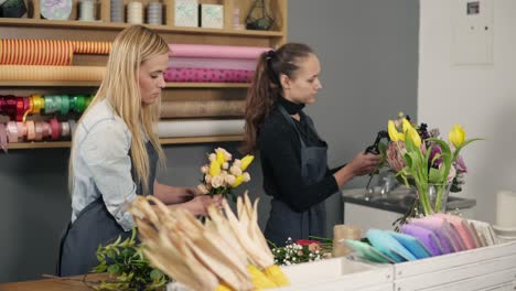 attractive blonde florist in apron standing with her coworker at counter in floral shot while arranging bunch of flowers