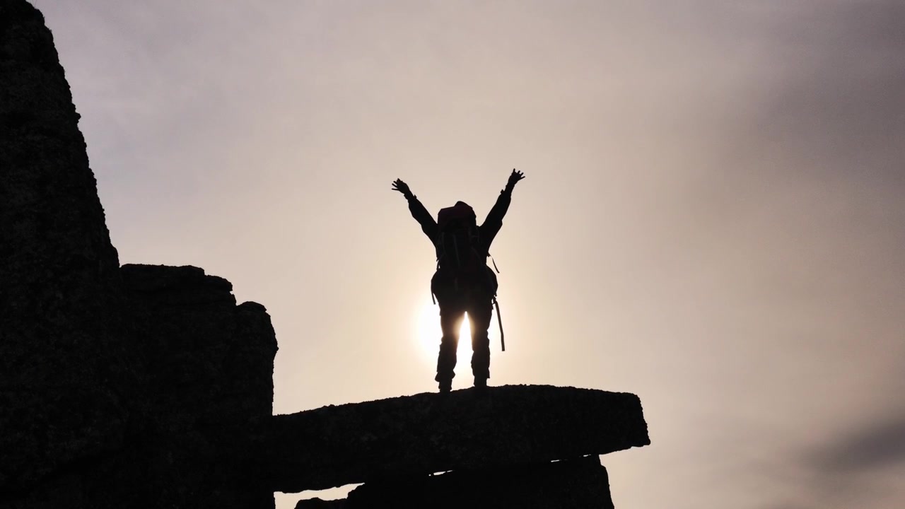 A silhouetted hiker wearing a backpack stands on a rocky outcrop, raising both arms upward in a wide gesture against a bright, overcast sky.